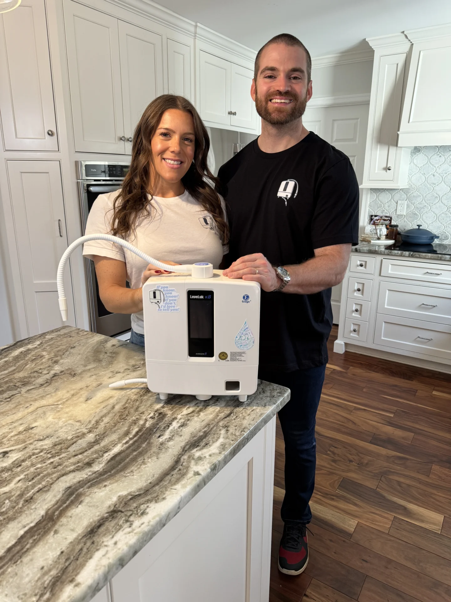 A man and woman smiling while standing in a kitchen, next to a white machine on the countertop.